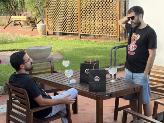Dos salseros conversando junto a una mesa de jardín con copas y bolsas de regalo negras.